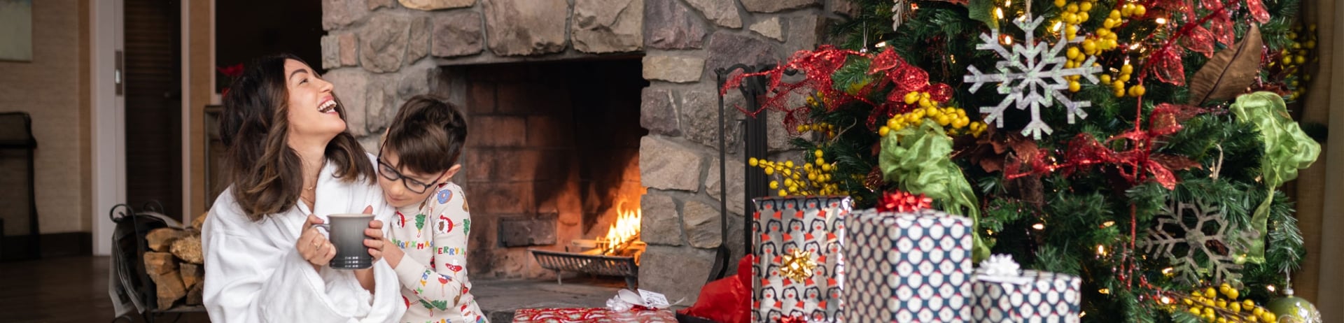 Mother and son smiling in front of the fireplace while sitting beside presents under the Christmas tree in Stanley Thompson Cabin at Fairmont Jasper Park Lodge.