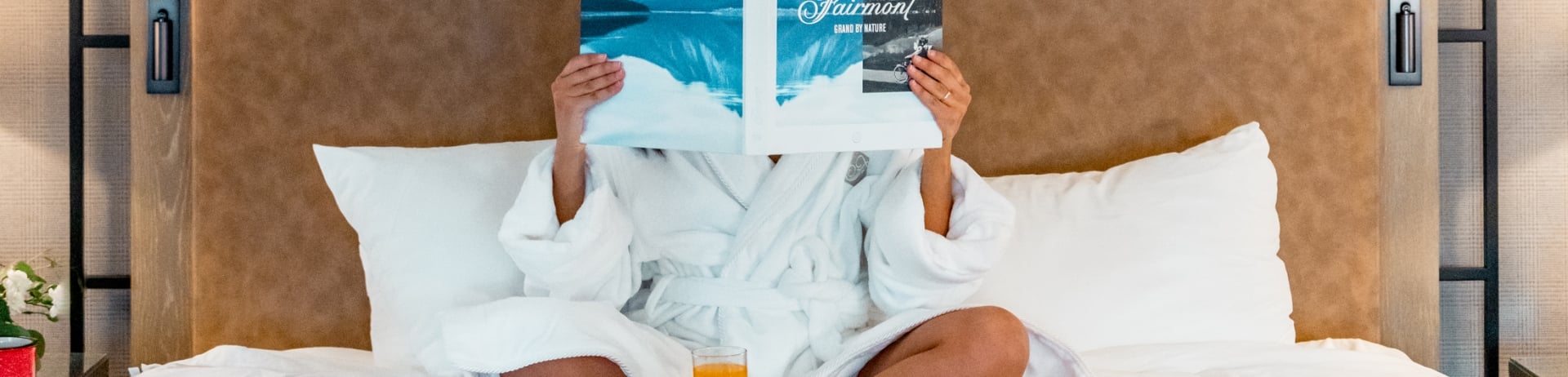 A woman sits on a bed reading a book while room service breakfast sits in front of her at Fairmont Jasper Park Lodge.