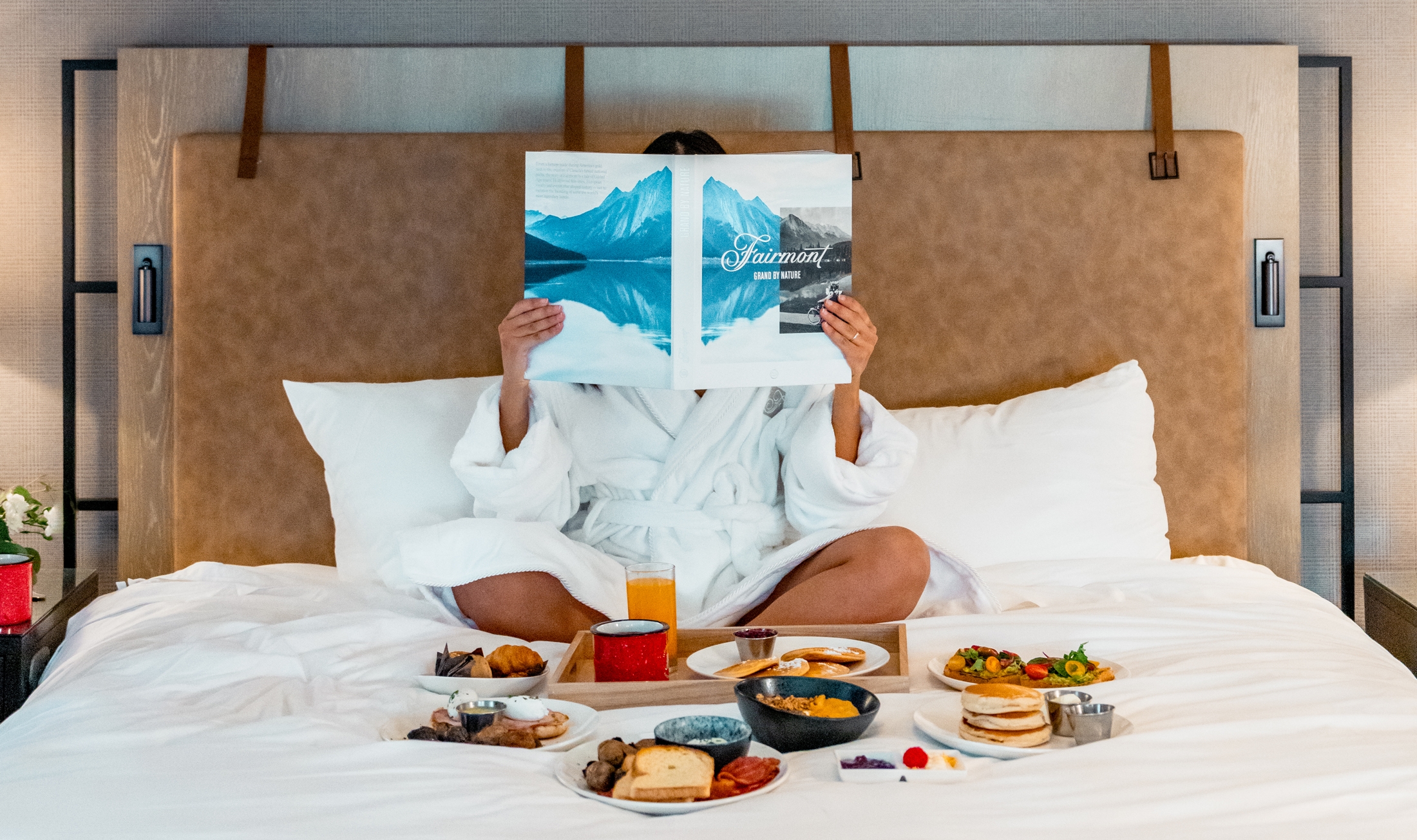 A woman sits on a bed reading a book while room service breakfast sits in front of her at Fairmont Jasper Park Lodge.