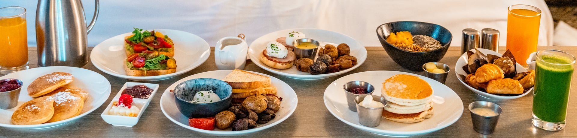 A woman lays on the bed reading a book while a lineup of room service breakfast items sit on the table waiting for her at Fairmont Jasper Park Lodge.