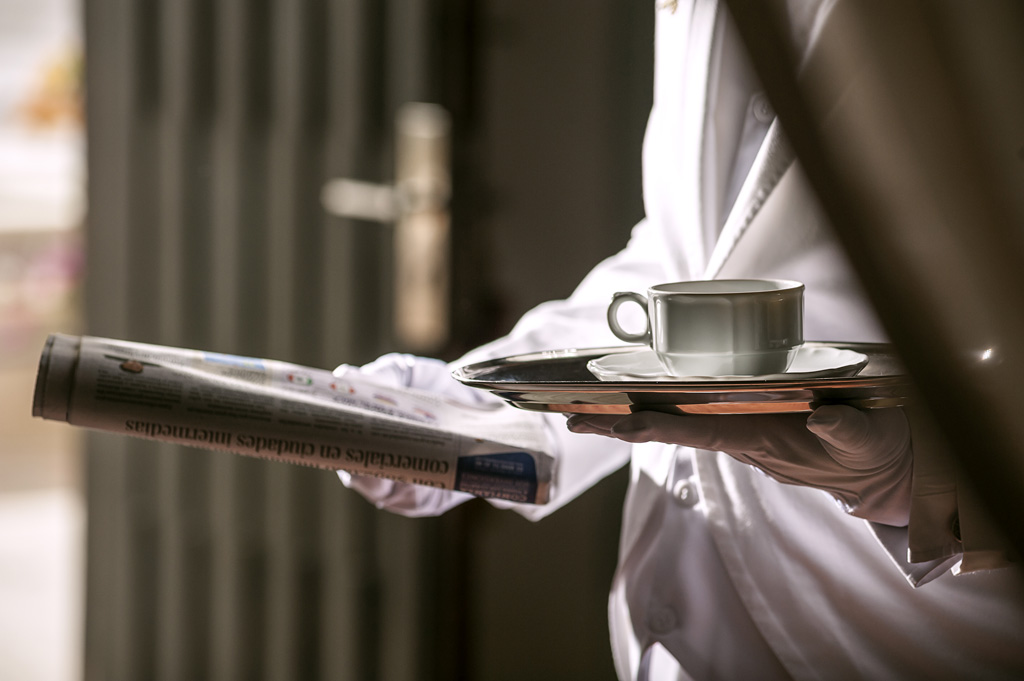 A butler dressed in white holding a newspaper and coffee