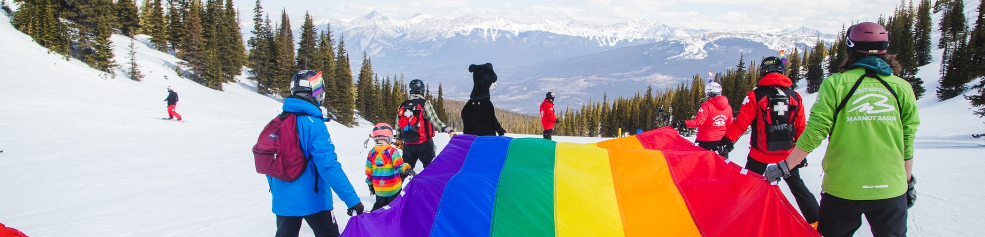 Skiers take a rainbow flag down a hill with mountains in the background at Jasper Pride Week