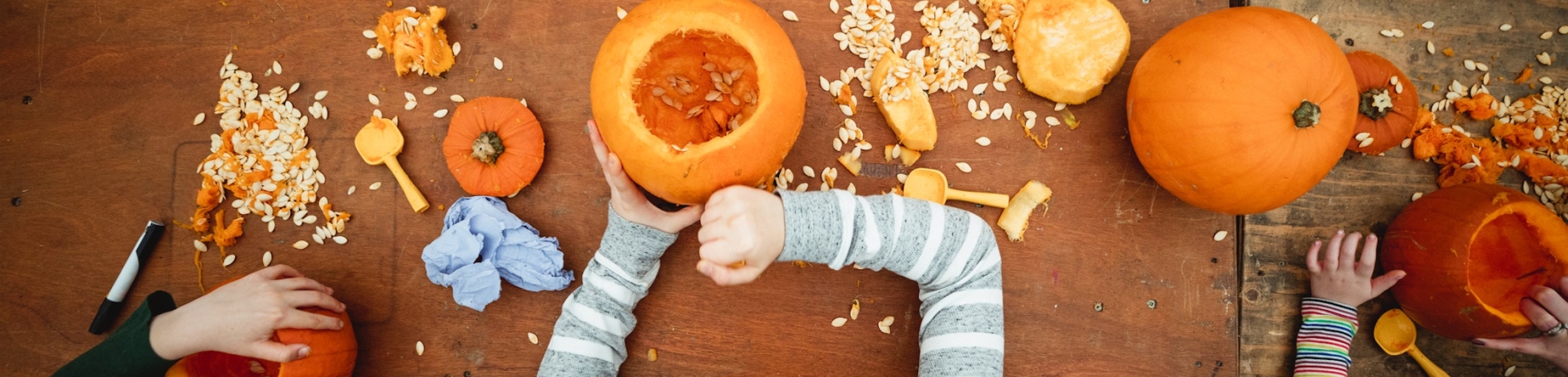 Direct above view of children siblings carving pumpkins at a farm after picking themin preparation for Halloween. There are pumpkin seeds scattered over the table and carving utensils.