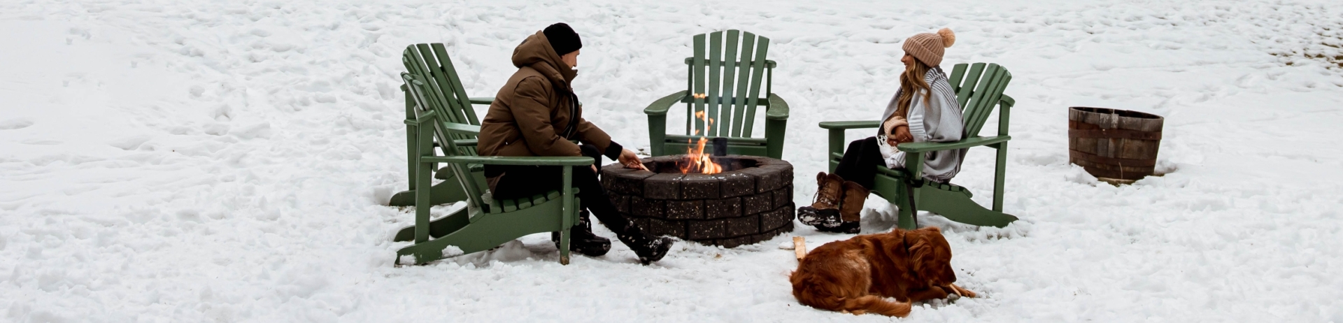 A couple sitting around a fire with their golden retriever in camp chairs, snow covers the ground in the yard of a Signature Cabin at Jasper Park Lodge