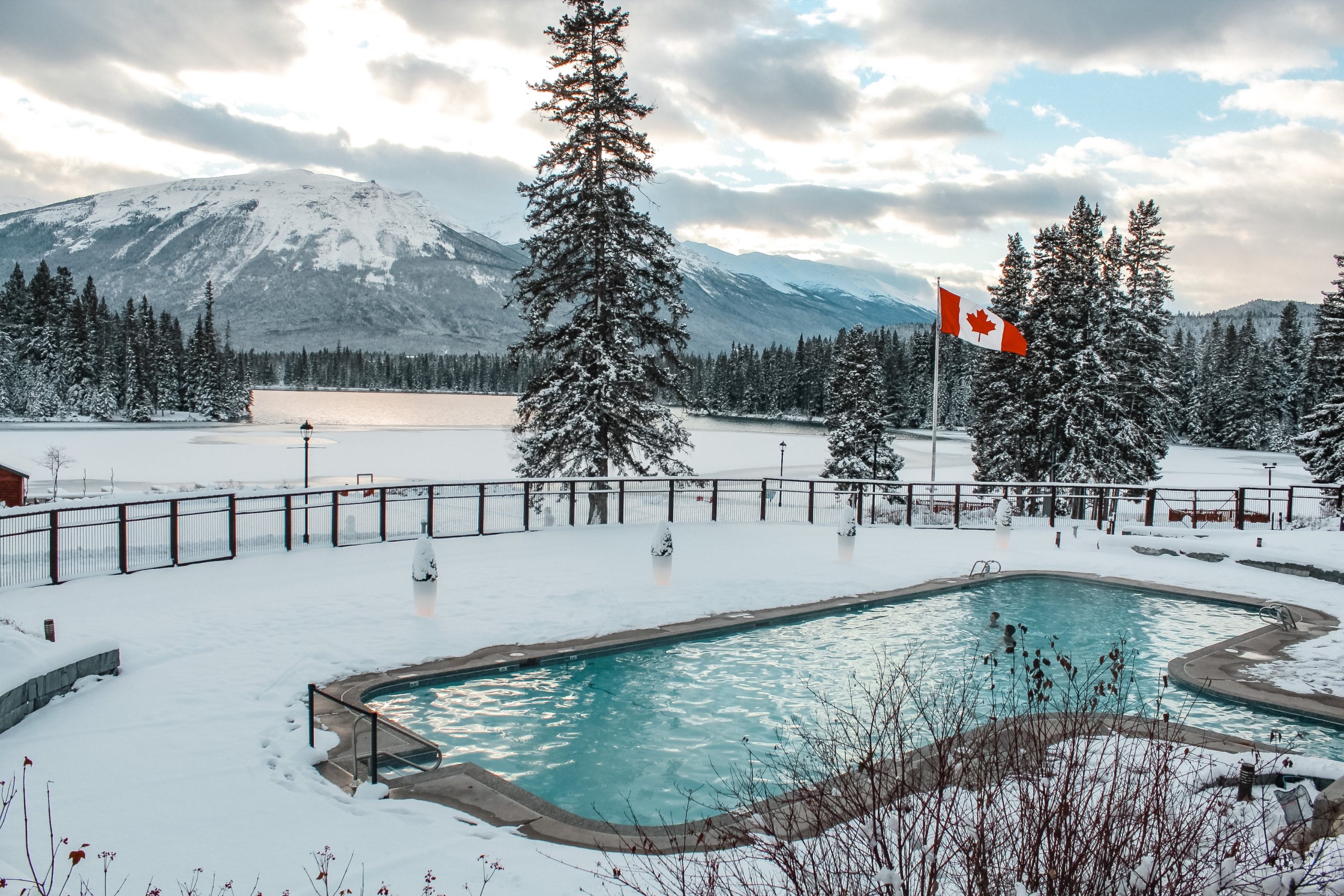 An outdoor pool overlooking the mountain range of Jasper Park Lodge in Jasper National Park. Snow covers the ground around the blue pool.