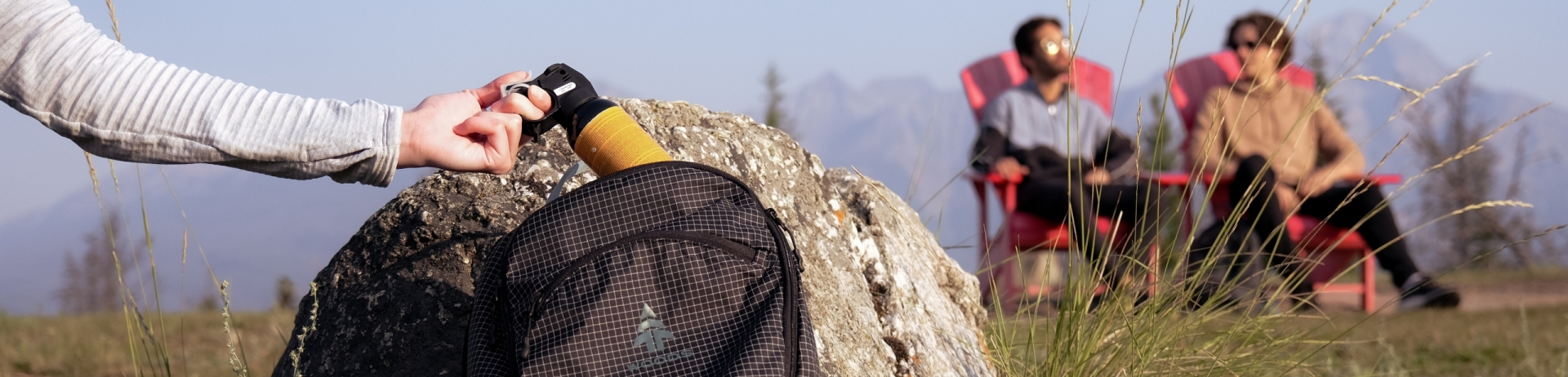 Hand removing bear spray from a hiking backpack while two hikers sit in chairs in the background.