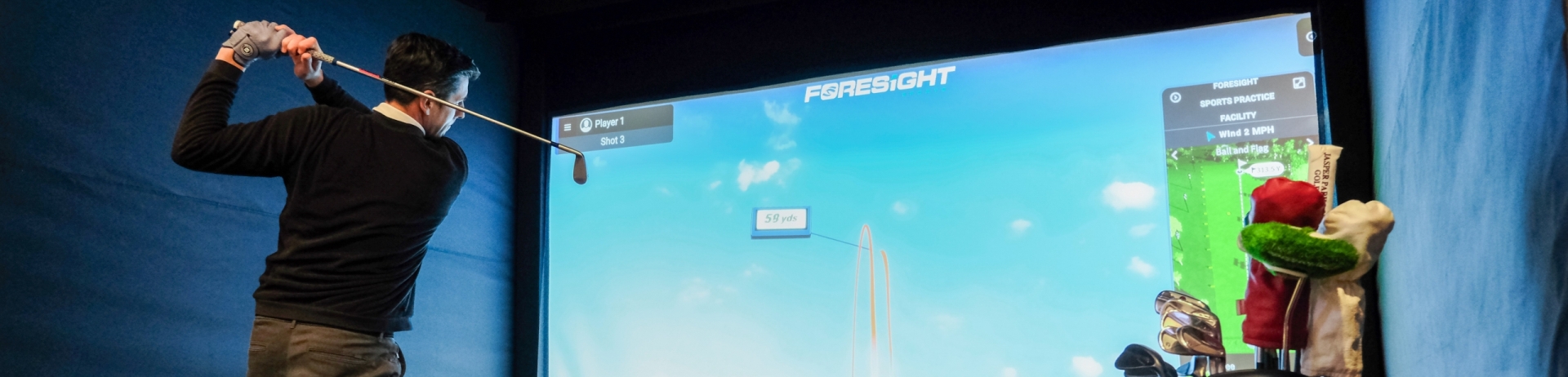 A man in athletic attire stands in front of a high-tech golf simulator, gripping a golf club as he prepares to take a swing. The simulator's vibrant screen displays a realistic golf course at Fairmont Jasper Park Lodge Golf Club.