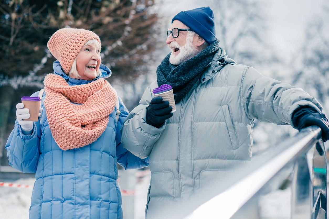 Positive senior man and woman with cups of coffee walking in winter day and looking at each other with a happy smile