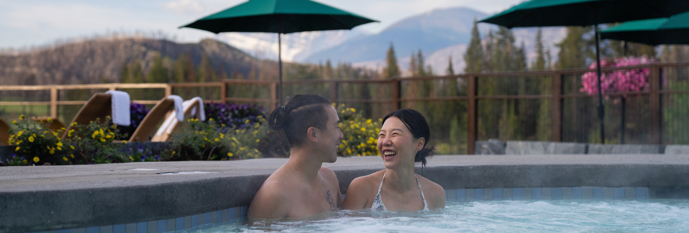Couple sitting in a hot pool with trees and mountain views behind them.