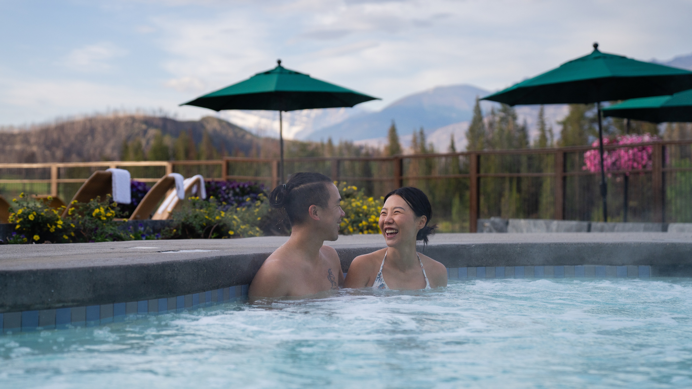 Couple sitting in a hot pool with trees and mountain views behind them.