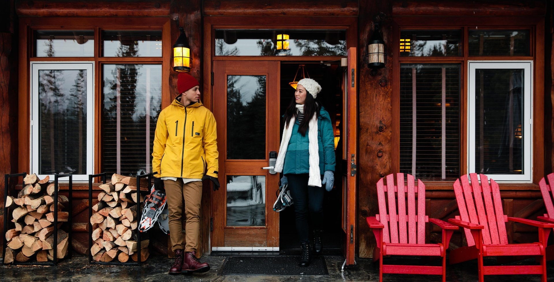 Couple standing outside a cabin in winter layers holding snowshoes