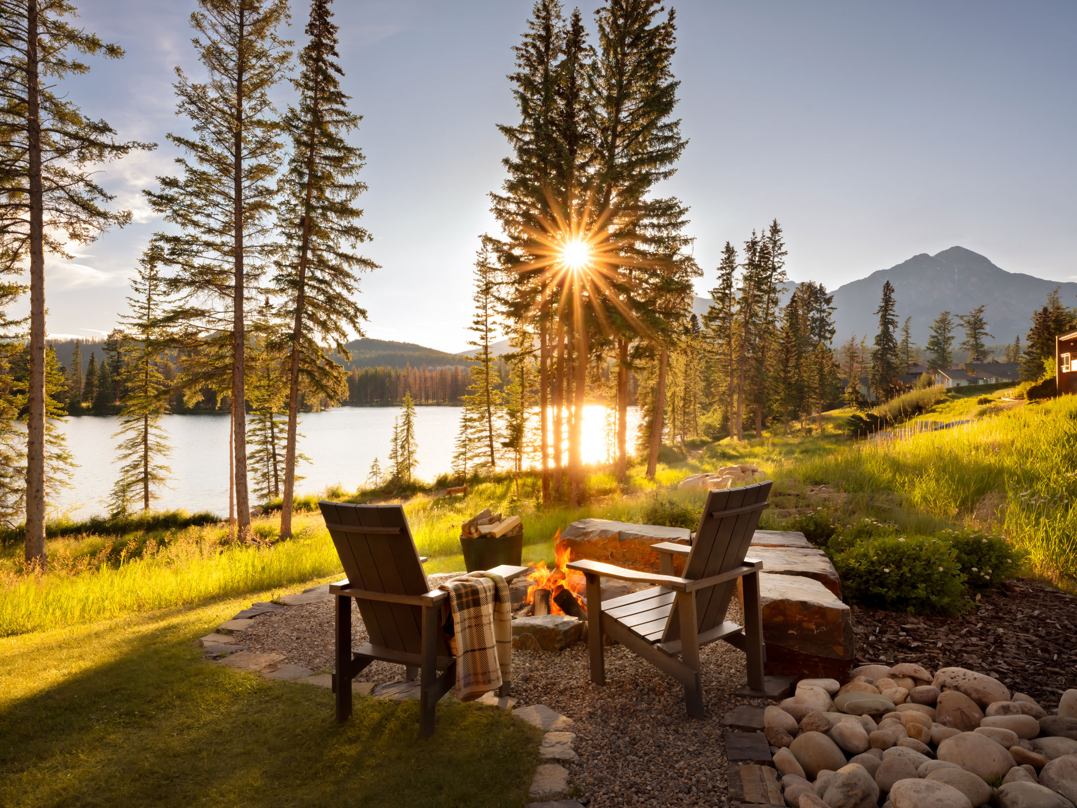 Autumn outdoor seating area at Fairmont Jasper Park Lodge overlooking Lac Beauvert, with Adirondack chairs, a stone fire pit, golden sunlight through the trees, and mountain views in the distance.