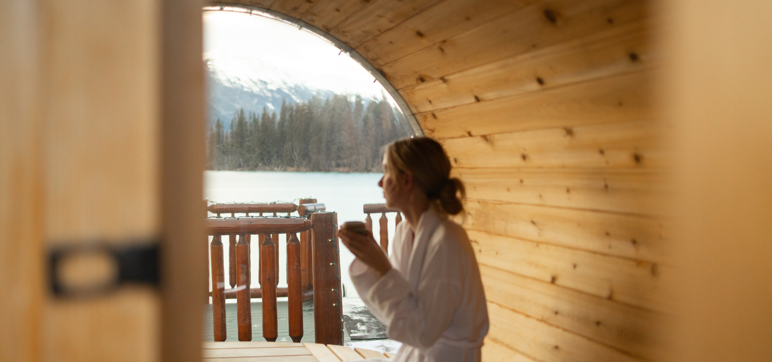 A woman warming her hands in a barrel sauna while looking out at the frozen lake.