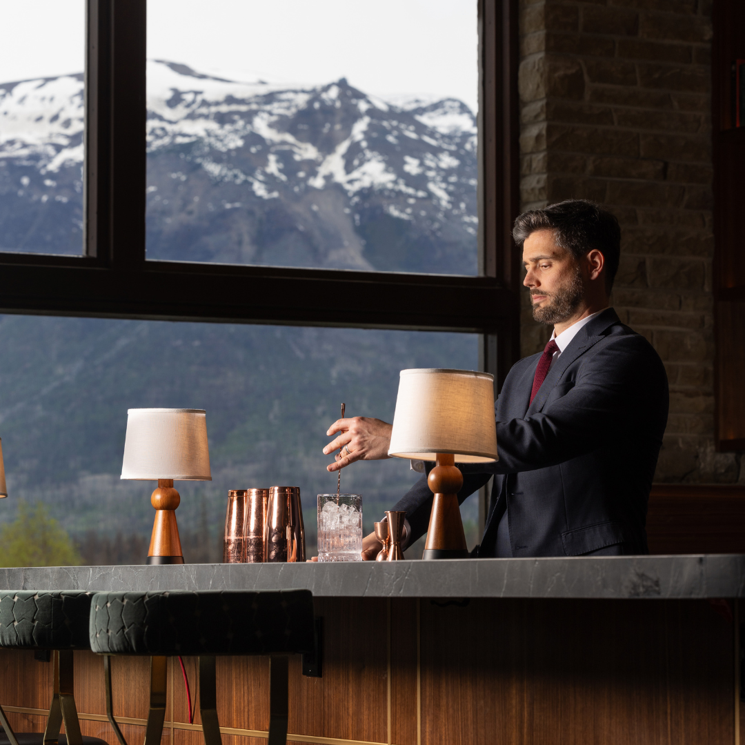 A bartender making a cocktail in front of a window with a mountain view.