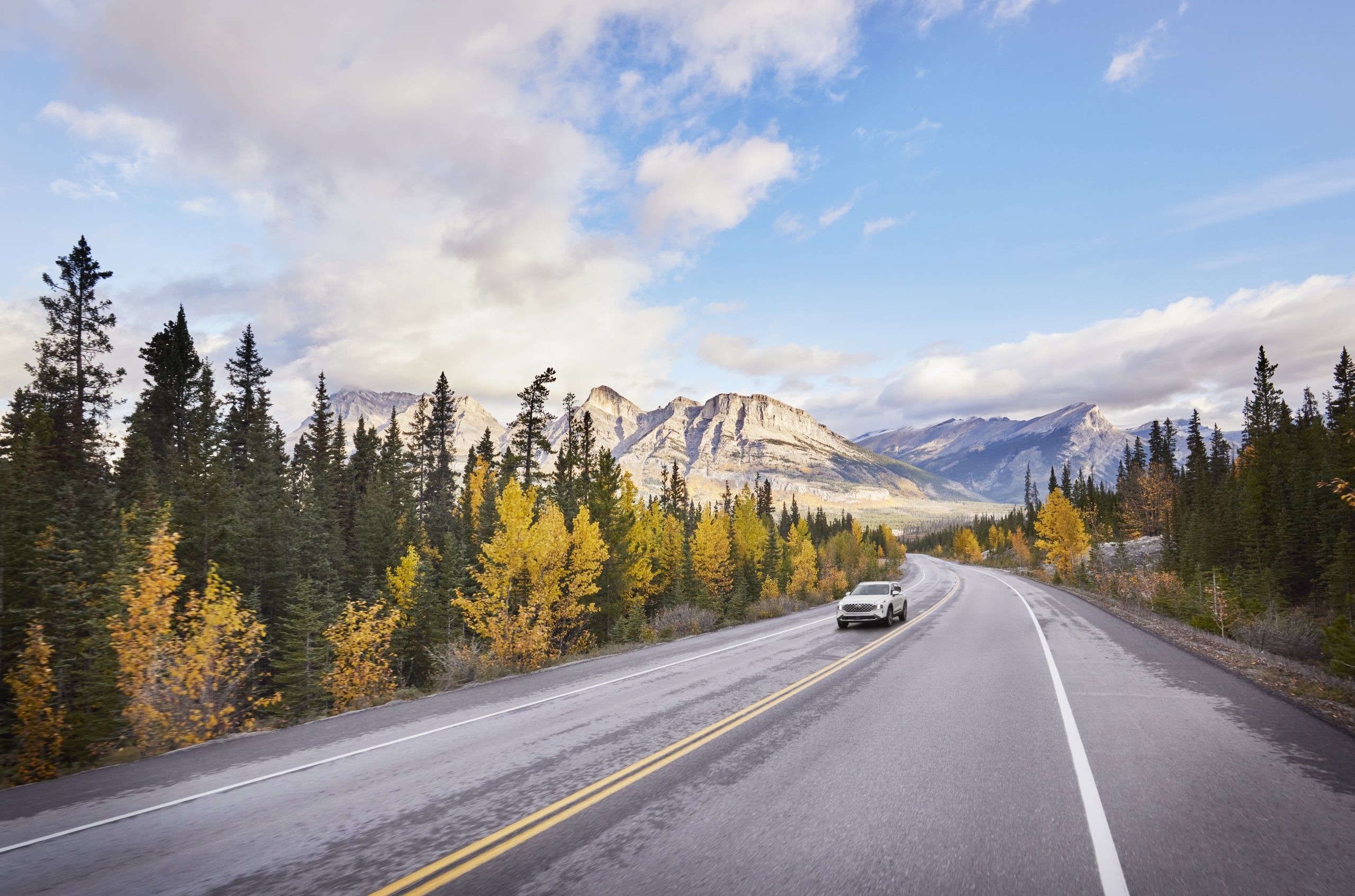 Car traveling down a highway with fall trees and mountains in the background.